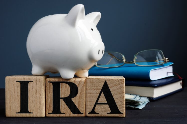 building blocks with the letters IRA on it, a piggy bank, dollar bills, books and glasses